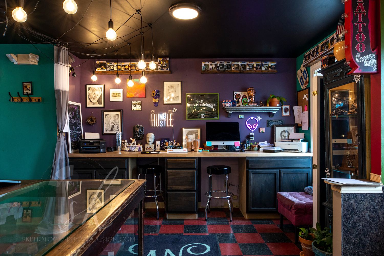 Eclectic tattoo shop interior with purple walls, dark wood counter, bar stools, and vintage art displayed under warm pendant lights.