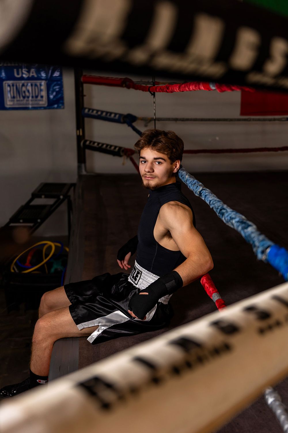 Boxer in black training gear leans against ropes in dimly lit boxing ring during intense workout session. - senior photos in Omaha, NE