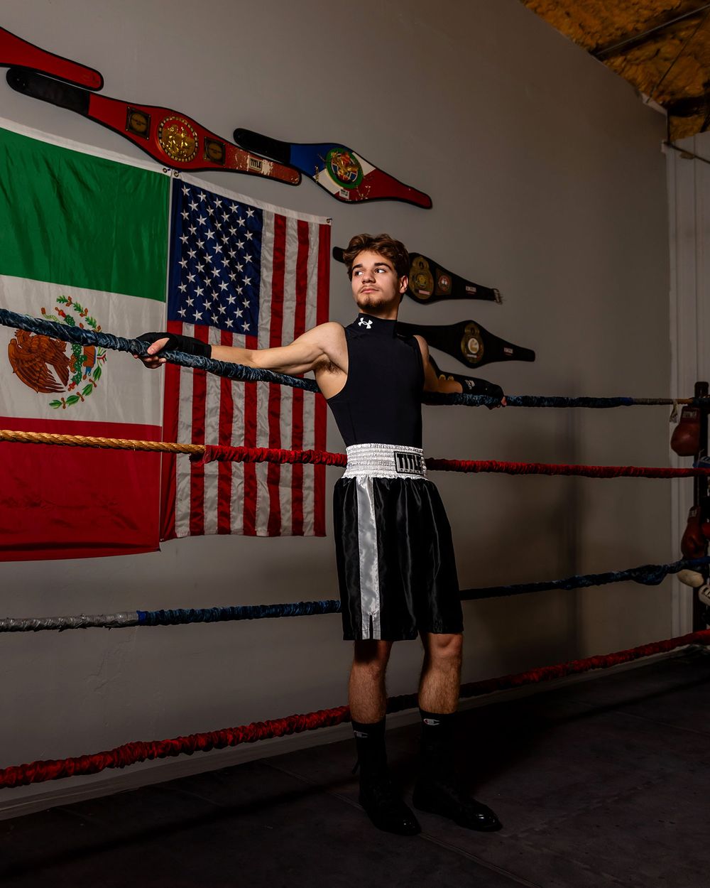 Athletic figure in black shirt and shorts performs boxing stance against wall with Mexican and American flags.  - senior photos in Omaha, NE