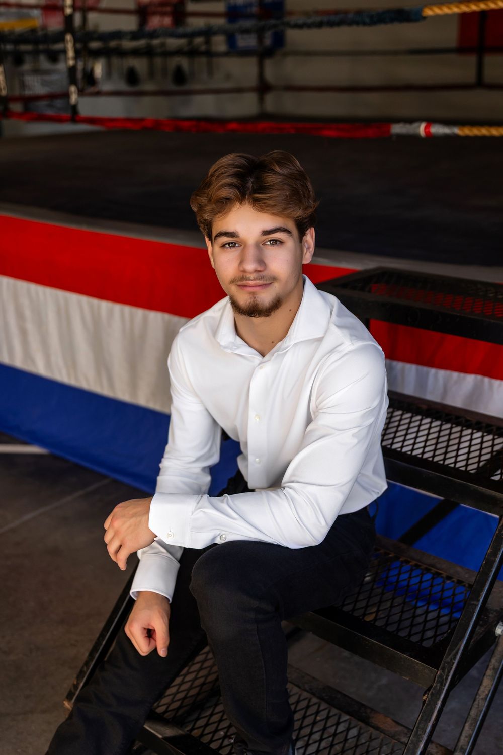 Young athlete in a white button-down shirt sits on bleachers inside a boxing gym with red and blue decorations. - senior photos in Omaha, NE
