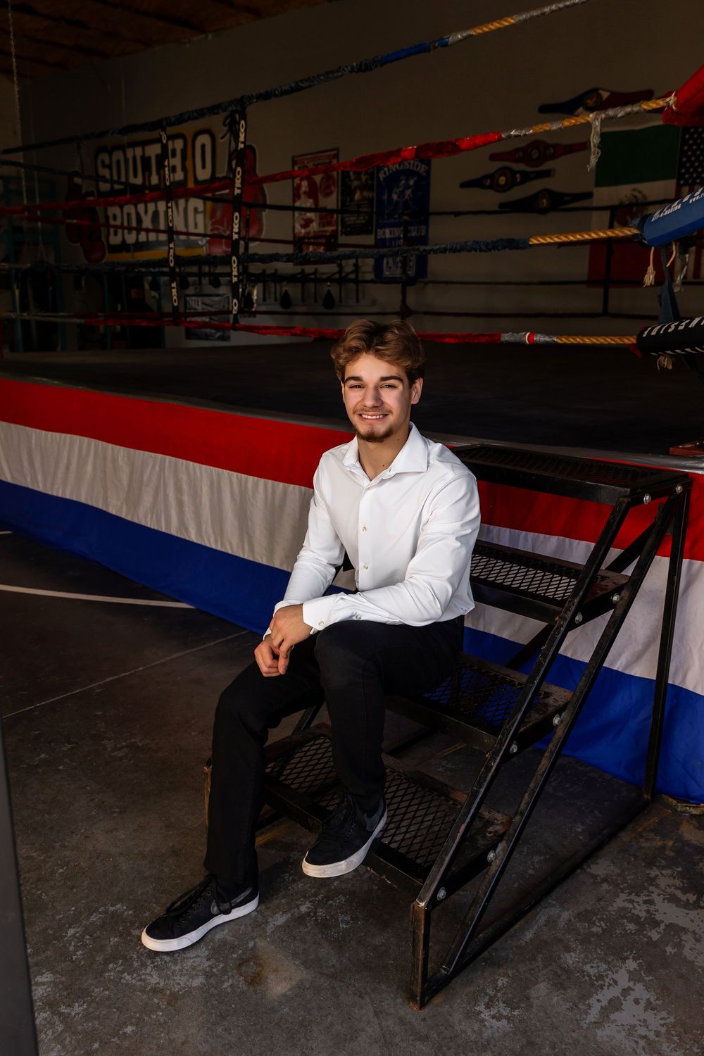 Person in white dress shirt and black pants sits casually on a dark bench against a wall with red and blue decor. - senior photos in Omaha