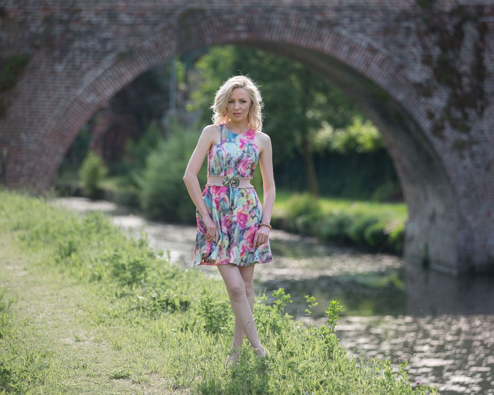 Stylish and fashionable young woman poses, framed against archway of bridge over river