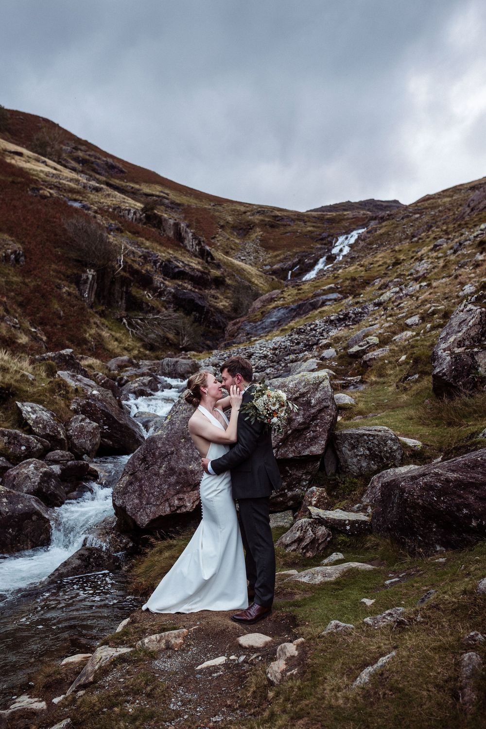 A couple shares a romantic moment by a cascading waterfall in the rugged Scottish Highlands on a moody, overcast day.