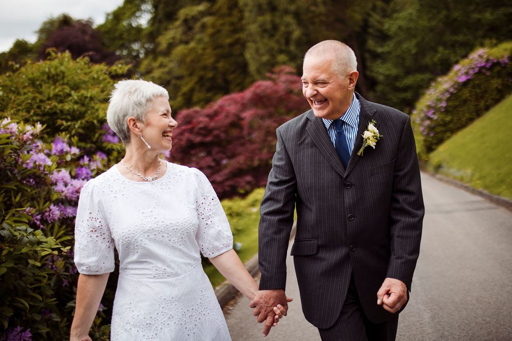 A couple in formal wedding attire walk and laugh together on a winding garden path surrounded by colorful flowers.