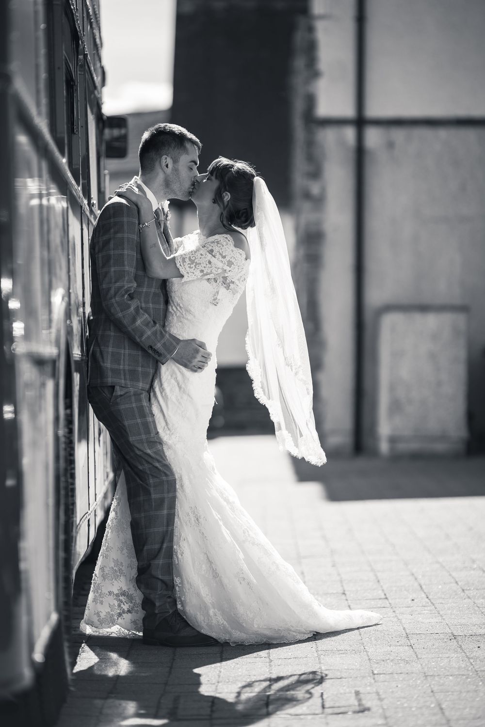 A romantic black and white wedding photo of a couple embracing against a wall, with the bride wearing a flowing lace gown and veil.