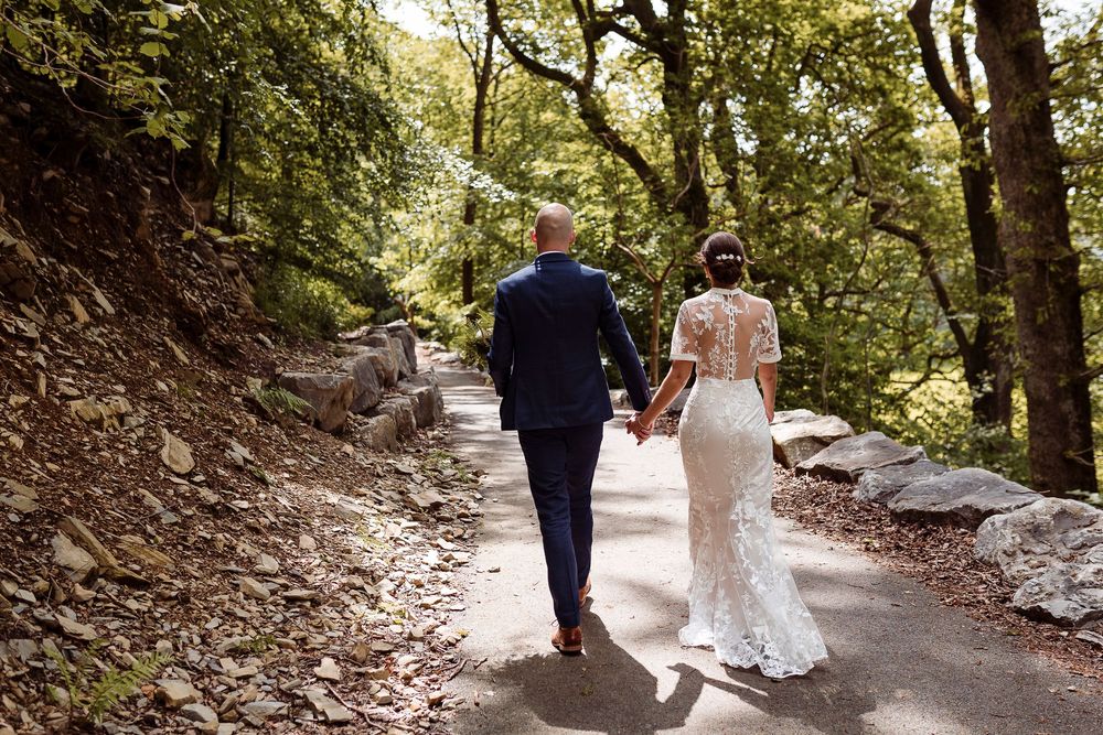 A couple walks hand in hand down a wooded forest path, with the bride wearing a fitted lace wedding dress.