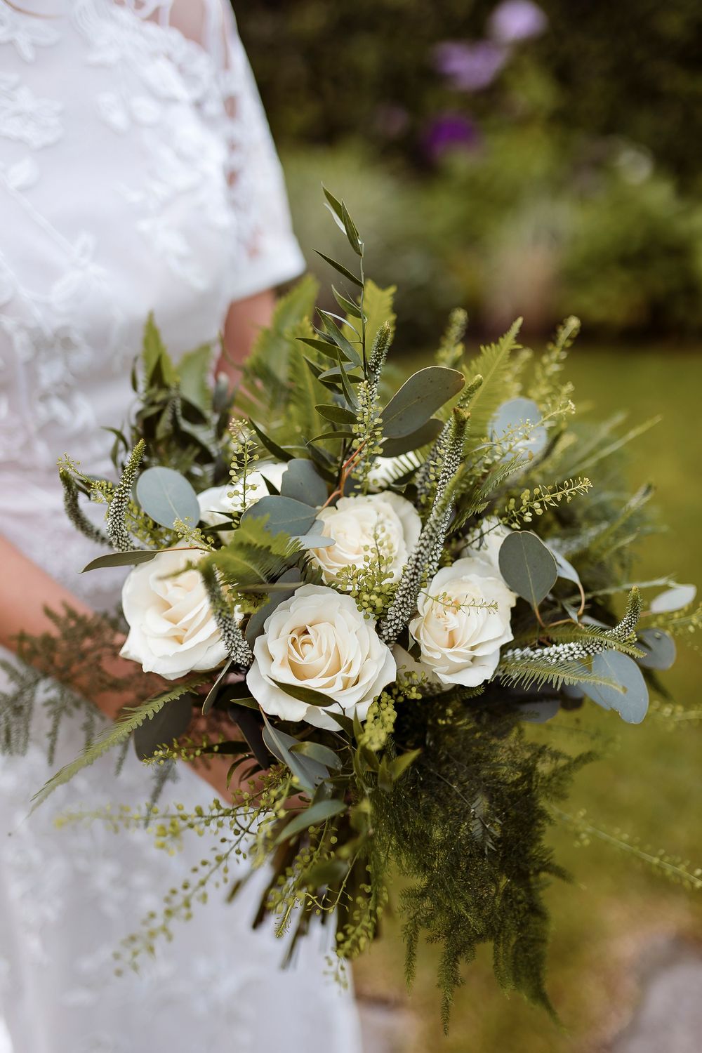 A romantic wedding bouquet with white roses, eucalyptus leaves, and delicate greenery against a lace dress.