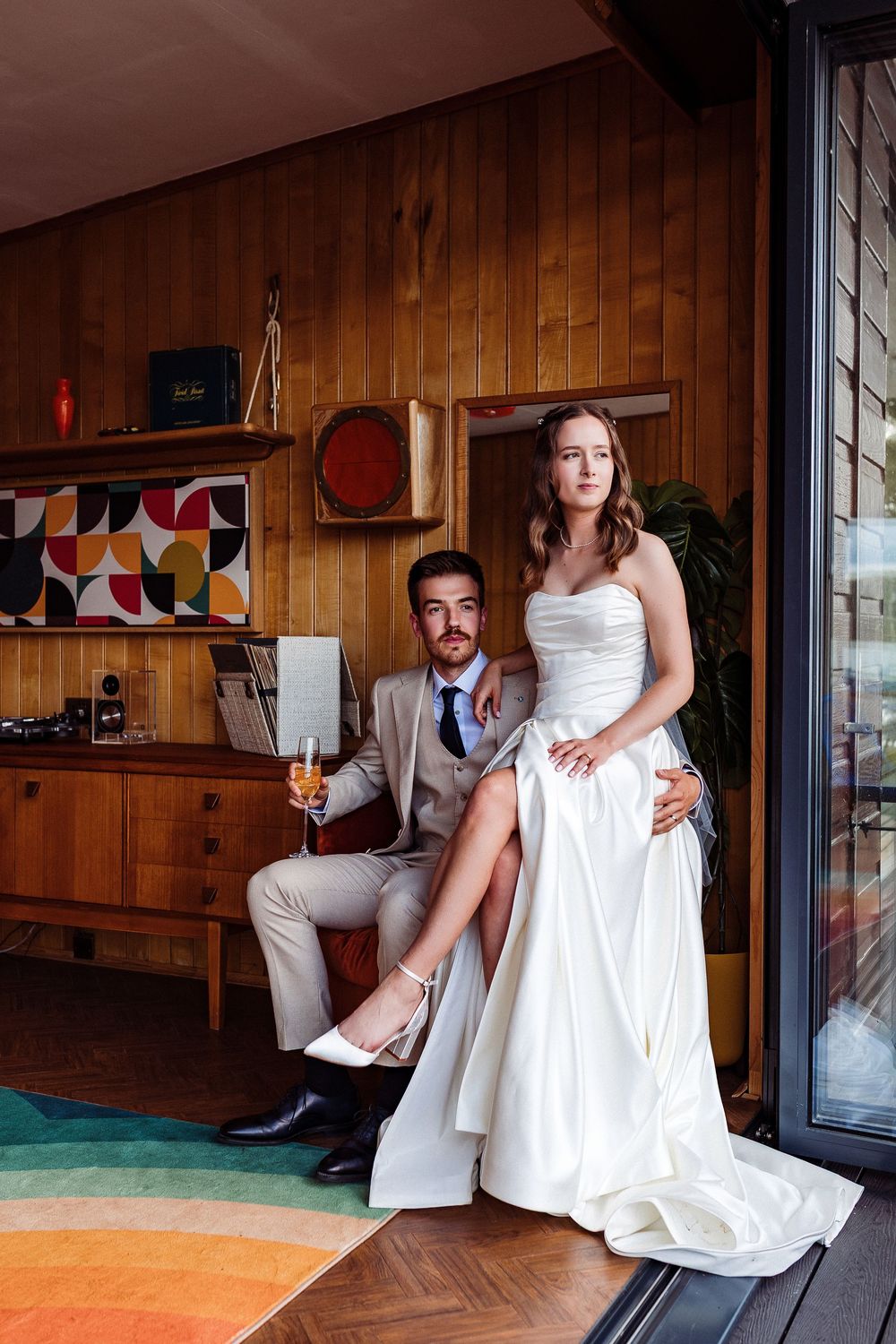 A couple in wedding attire poses together in a mid-century modern room with wood paneling and a colorful round rug.