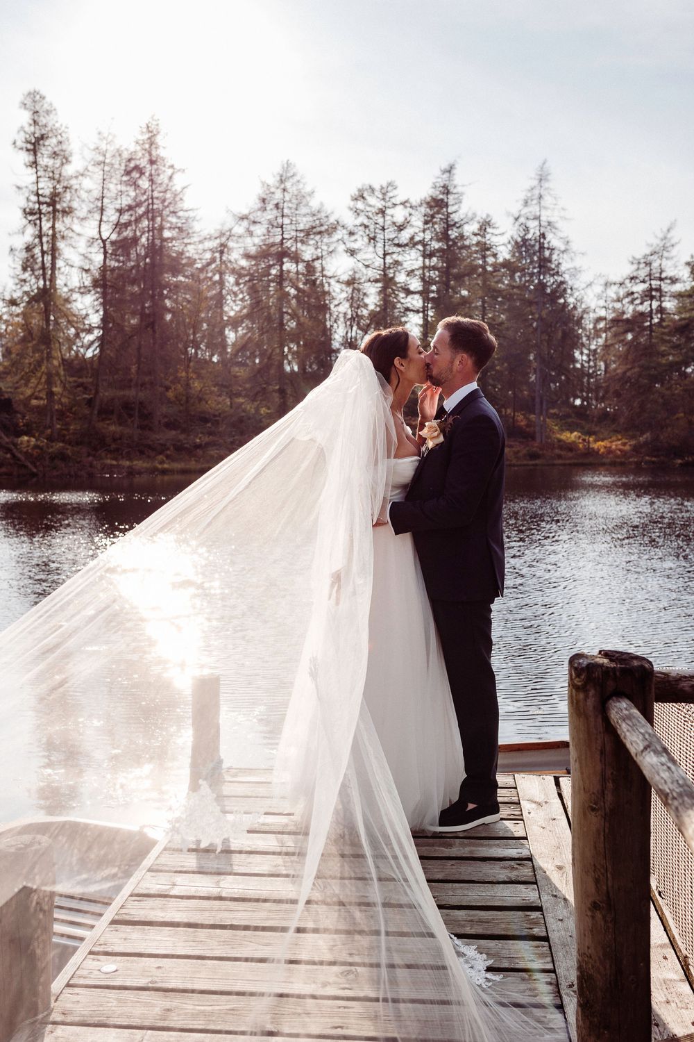 A romantic wedding couple shares a kiss on a wooden dock by a serene lake with cypress trees in the background.
