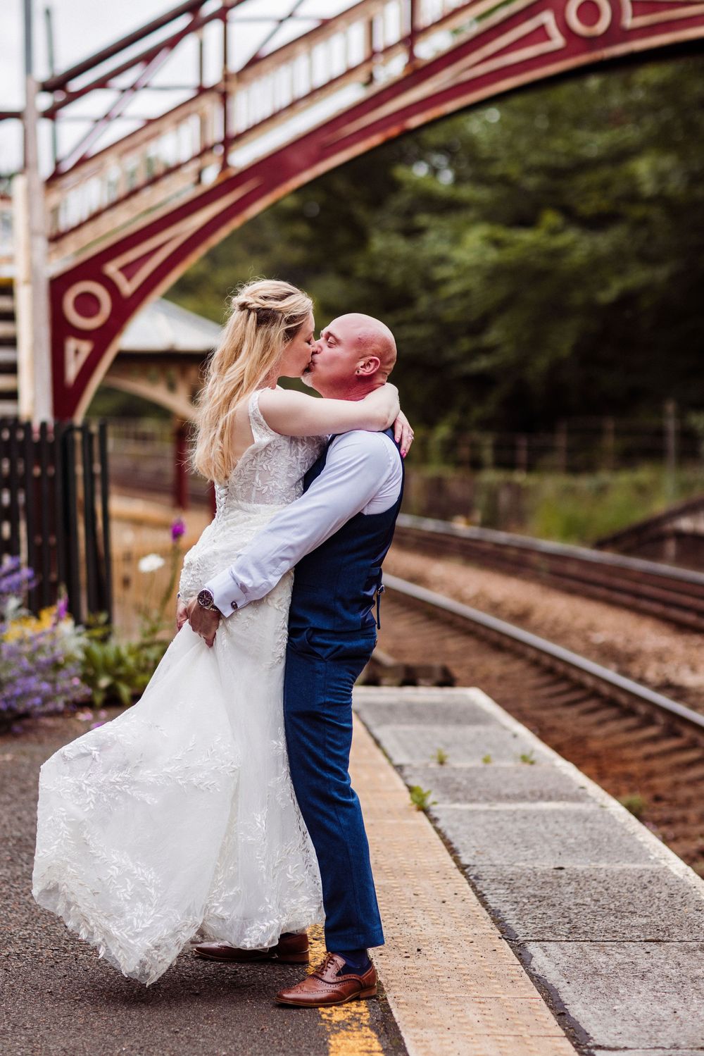 A romantic couple in wedding attire shares intimate moments at a train station platform.