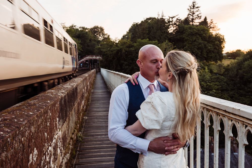 A romantic couple embraces on a stone bridge as a train passes by during sunset.