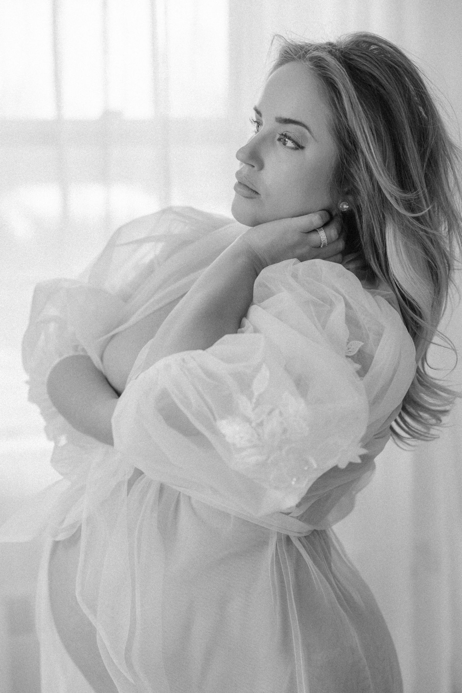 Woman in cream sweater dress reclines elegantly on white tufted sofa in bright sunlit room with sheer curtains.