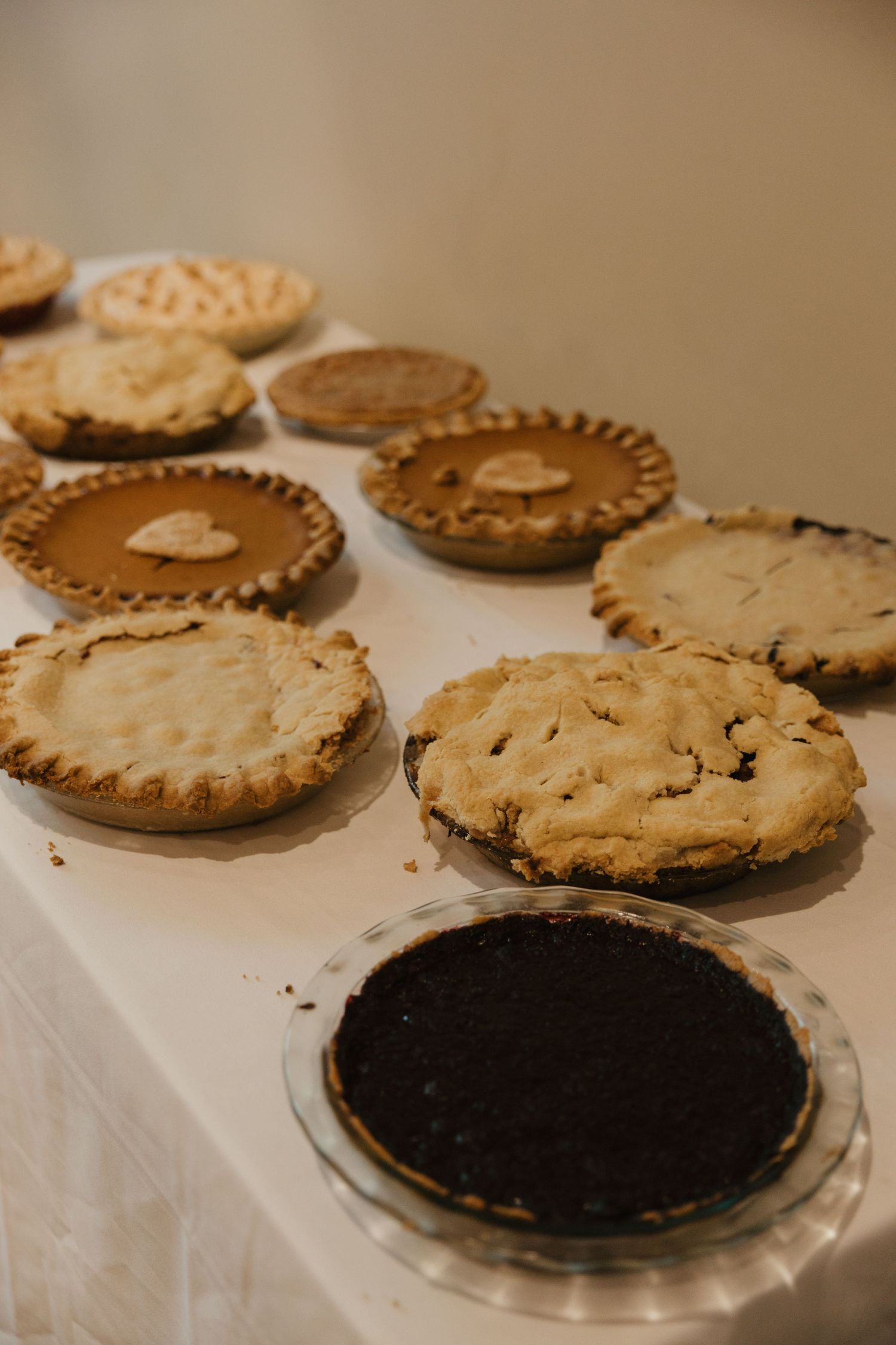 Assorted homemade pies displayed on a white tablecloth including pumpkin, fruit, and chocolate cream varieties.