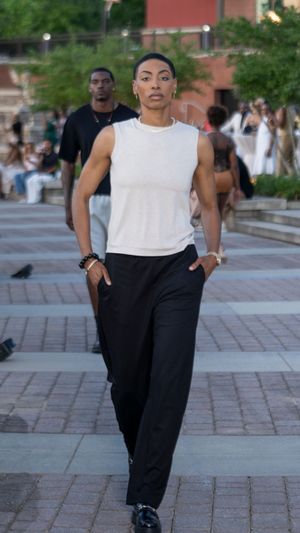 Model walks runway in white sleeveless top and black pants on brick courtyard.
