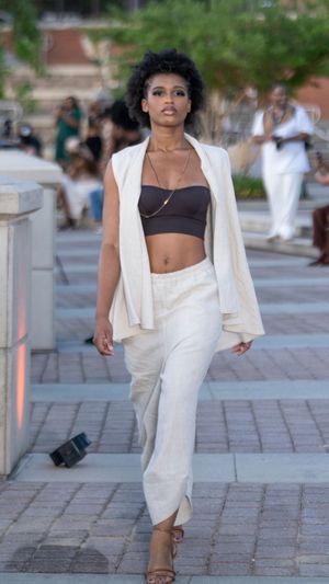 Fashion model walks down urban pathway wearing white suit and brown crop top during outdoor photoshoot.