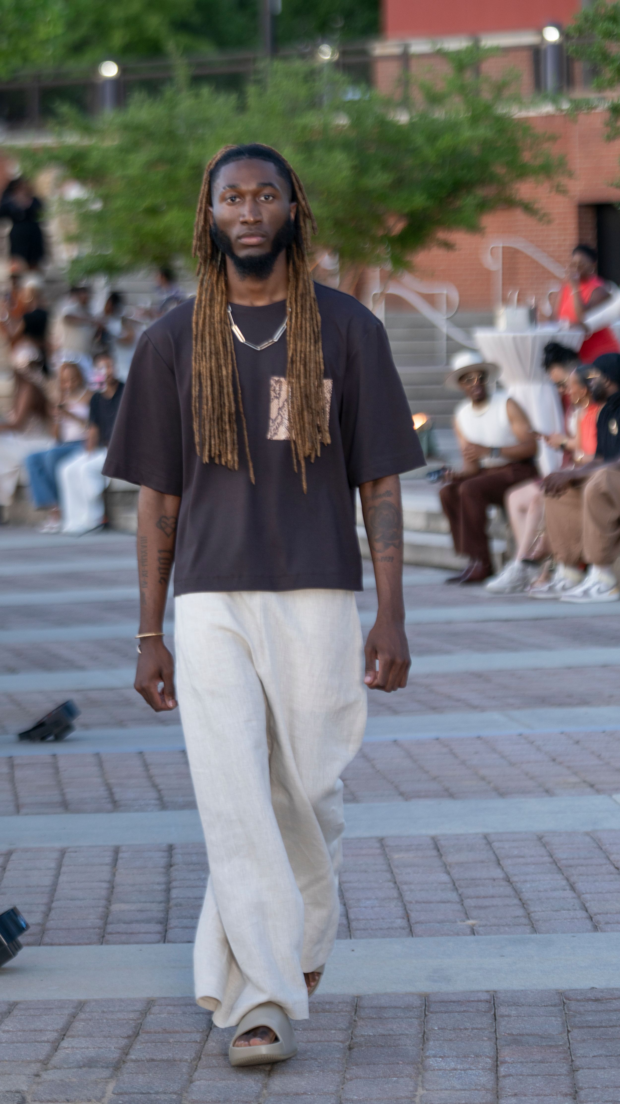 Model walks runway wearing black t-shirt and white wide-leg pants with long dreadlocks hairstyle.