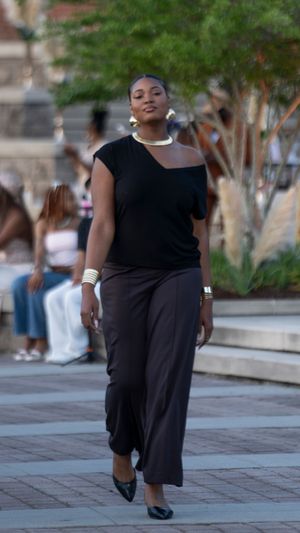 A model presents a black outfit while walking on a brick-paved outdoor runway.