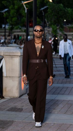 Person wearing brown suit and sunglasses walks confidently on stone pathway.