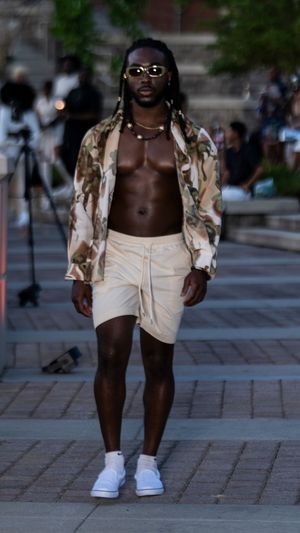 Model in camouflage jacket and cream shorts walking on outdoor runway.
