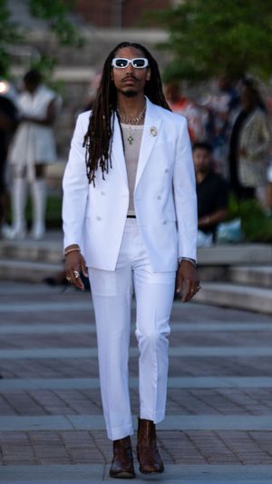 All-white suit ensemble accessorized with white sunglasses and brown boots on outdoor runway.