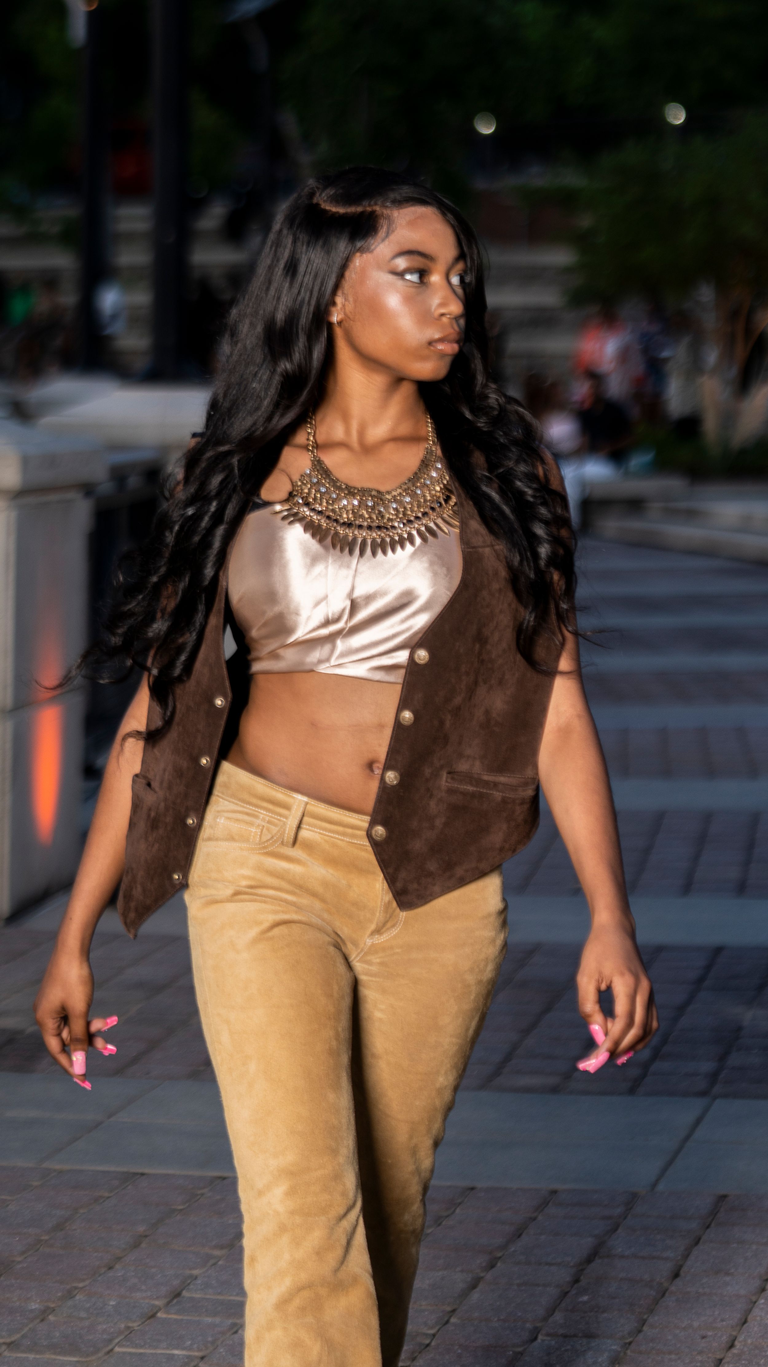 Model walks runway in brown vest, metallic crop top and tan suede pants.