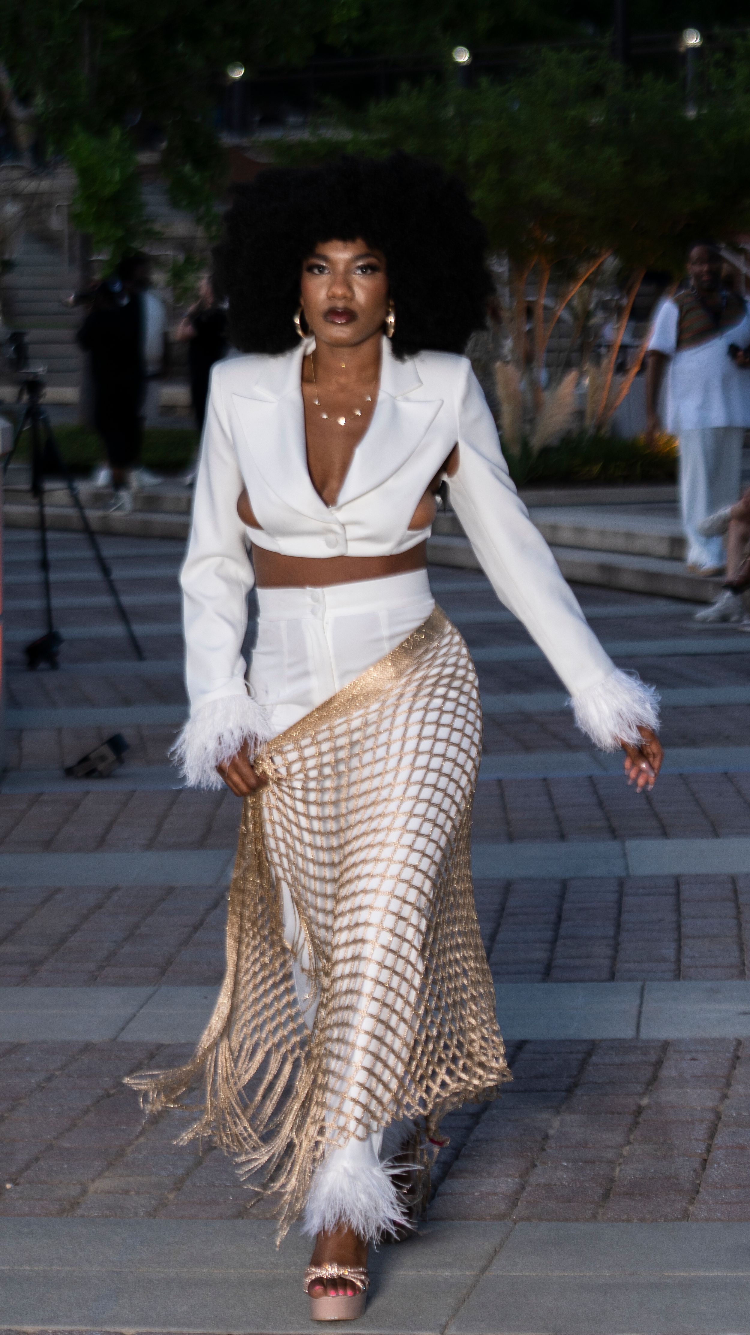 Model poses in white blazer and metallic sequined pants during outdoor fashion show.