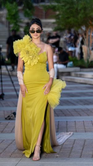 Model showcases dramatic yellow tulle gown with ruffled shoulder detail on outdoor runway.