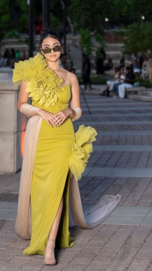 Elegant yellow one-shoulder evening gown with dramatic floral detail and side slit on stone paved walkway.