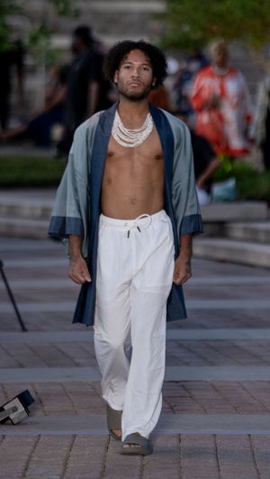 A shirtless person in white pants and gray jacket walks on a brick paved street in an urban setting.