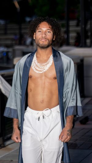 Model walks runway in open grey robe and white pants with layered pearl necklaces at outdoor fashion show.