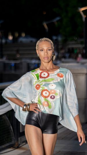 Fashion model walks runway in floral print asymmetrical top with black shorts during outdoor evening show.