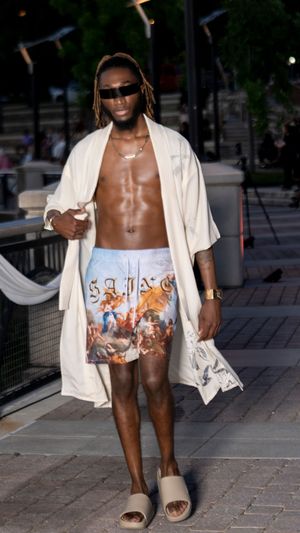 Model wearing artistic printed shorts and white robe walks runway in casual slides.