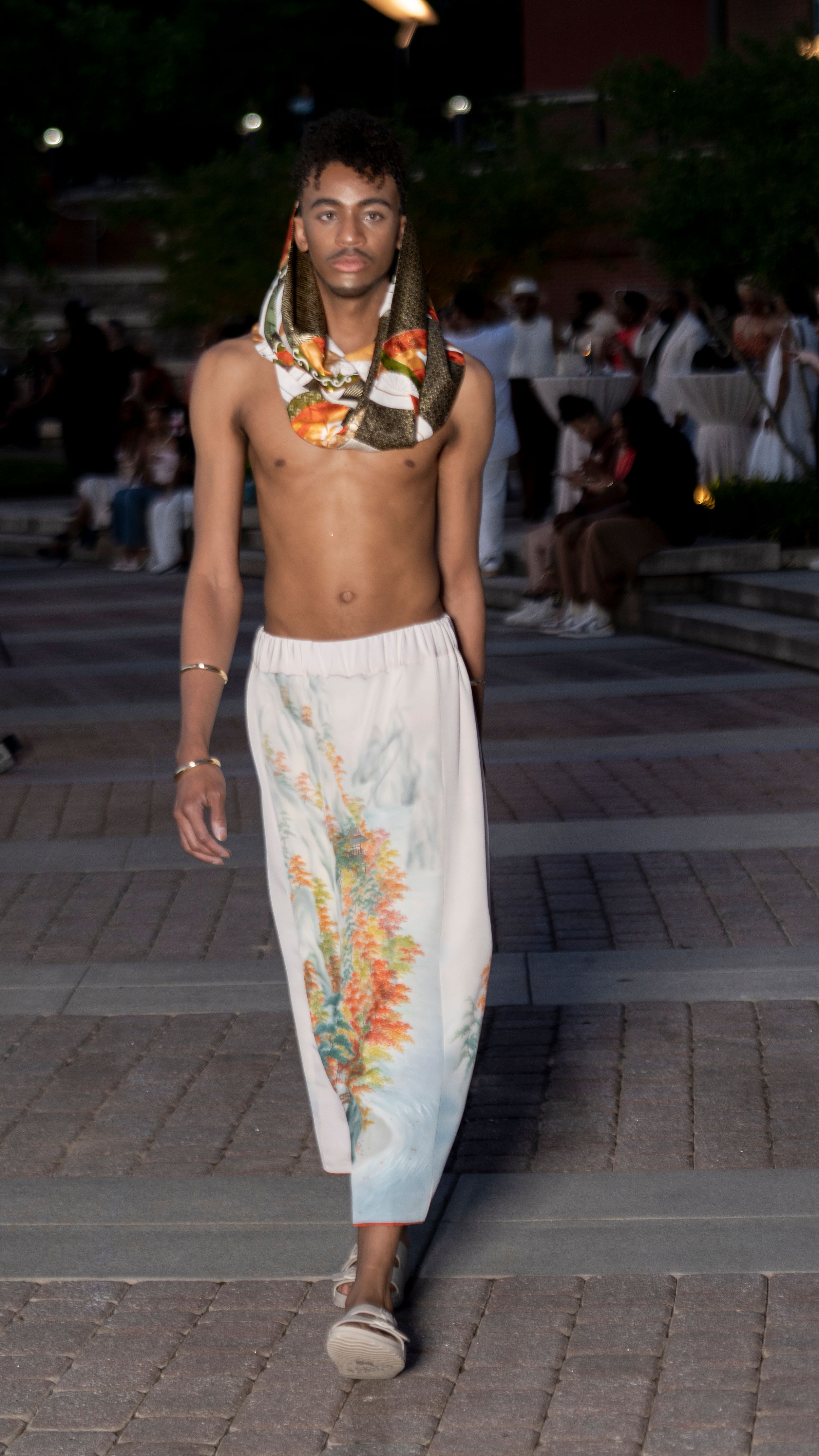 Fashion model wearing a white floral painted dress on a brick paved walkway at dusk.