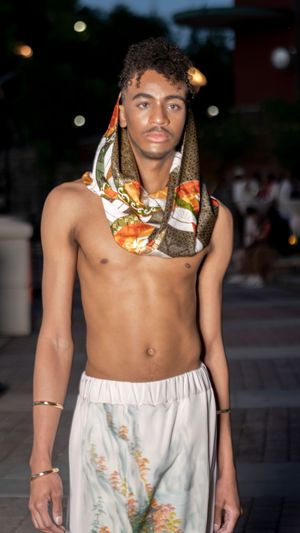Model in white flowing pants and patterned scarf poses outdoors at dusk.