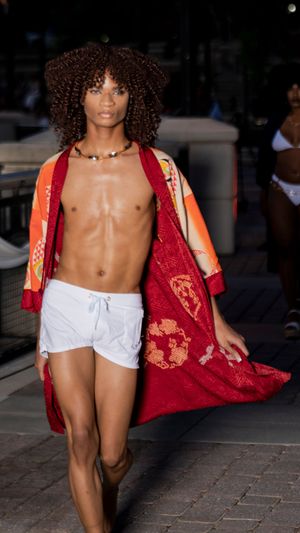 Model displays white shorts and red patterned robe on brick pathway during fashion show.