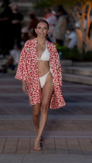 Model walks runway in red floral print jacket and white swimsuit during outdoor fashion show.