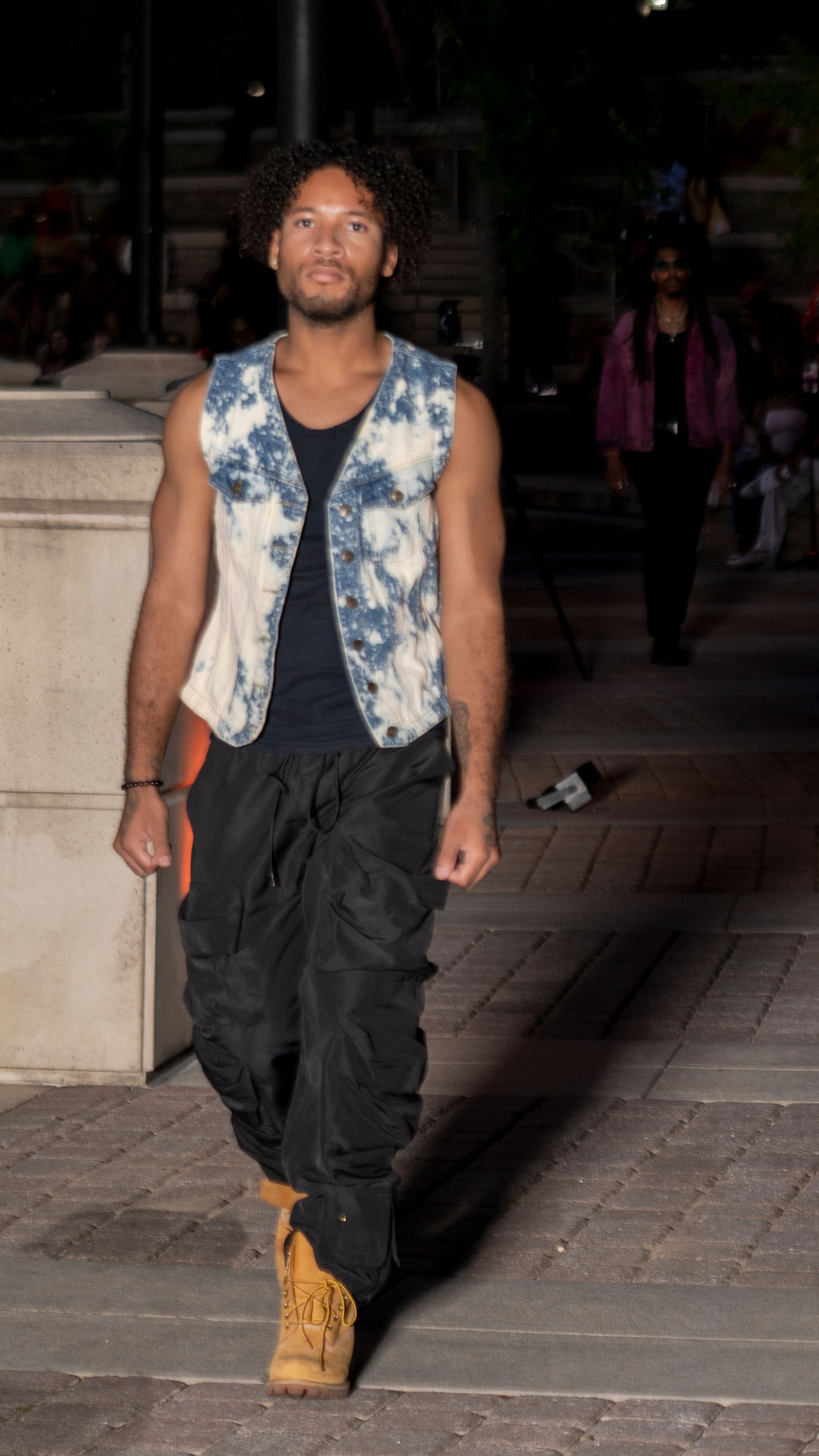 Person wearing tie-dye vest and dark pants poses on stone steps during evening photoshoot.