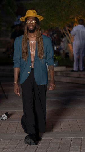 Runway model wearing teal blazer, black pants and straw hat walks during evening fashion show.
