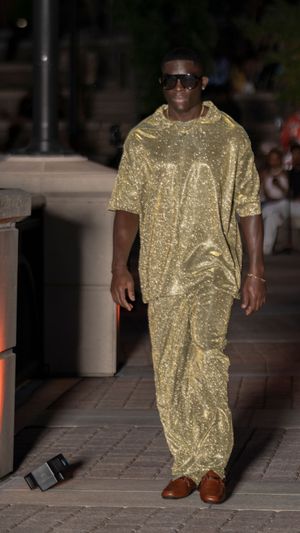 A model walks the runway wearing a shimmering gold oversized t-shirt dress and brown shoes.