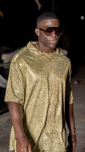 Person wearing shimmering gold button-up shirt and sunglasses walks on outdoor pavement.