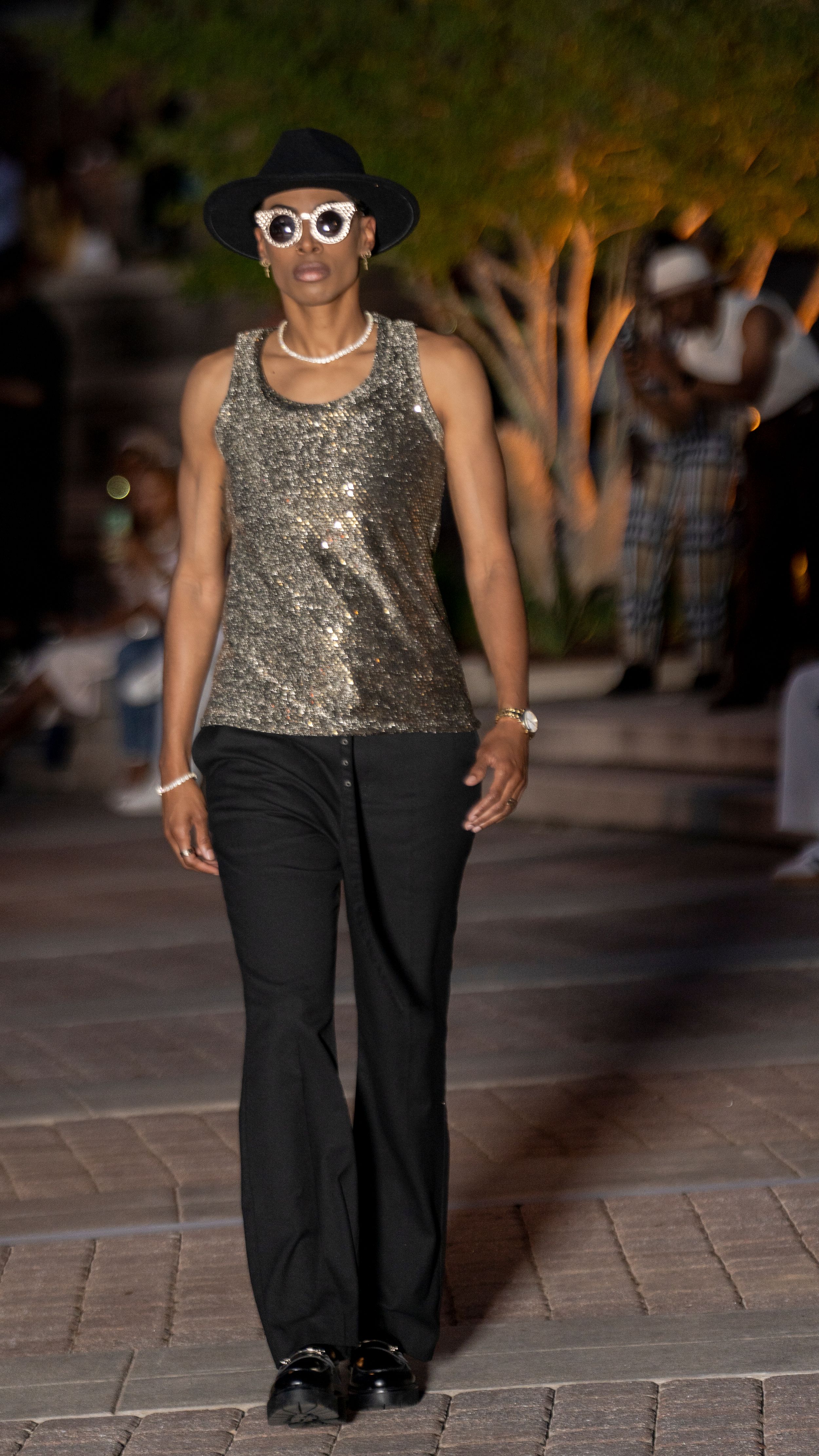 Model walks runway in sparkly silver sleeveless top and black pants at outdoor fashion show.