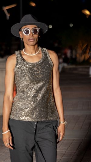 Fashion model wearing sequined tank top with black trousers and sunglasses on outdoor catwalk.