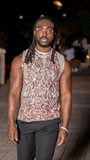 Model wearing patterned sleeveless top and black pants walks confidently on outdoor runway.