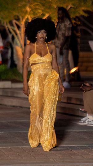 Model showcases flowing gold sequined gown on outdoor runway at dusk.