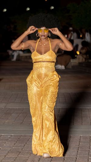 Model walks runway in sparkling gold evening gown with matching accessories at night.
