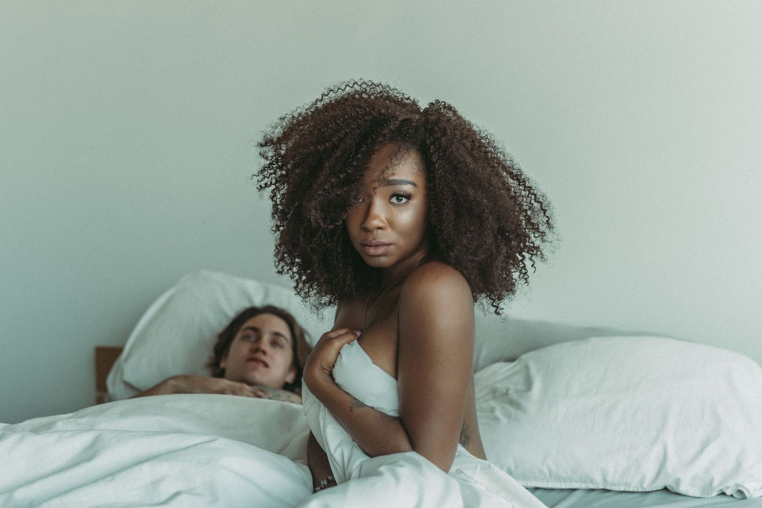 A couple relaxes together in bed with white bedding against a mint green wall.