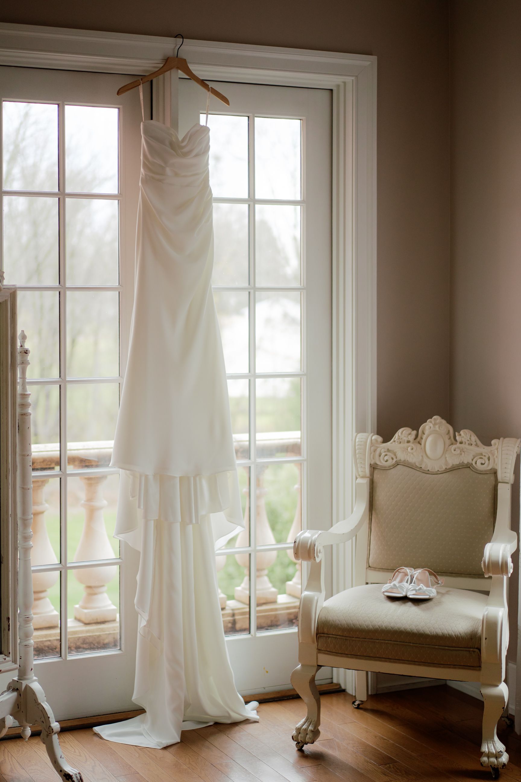 White wedding dress hanging in sunlit room with ornate vintage furniture.