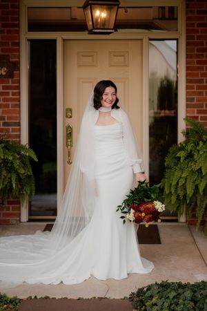 Bride in elegant white gown and veil posing on brick porch with hanging ferns and lantern.