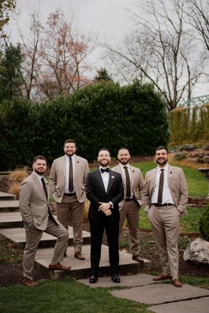 Groomsmen in tan suits pose casually on stone steps outdoors.