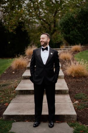 Two people in formal black tuxedos stand on a stone pathway surrounded by autumn foliage.